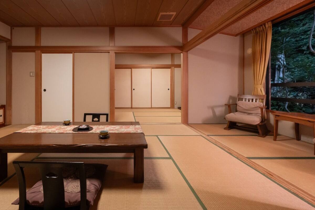 A traditional Japanese room with tatami flooring, a low table with a tea set, a sliding door, and a view of greenery outside.