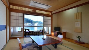 Um quarto japonês tradicional com tatames, uma mesa baixa, cadeiras, uma televisão e vista para a água.