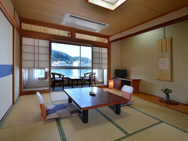 A traditional Japanese room with tatami flooring, a low table, chairs, a television, and a view of a body of water.