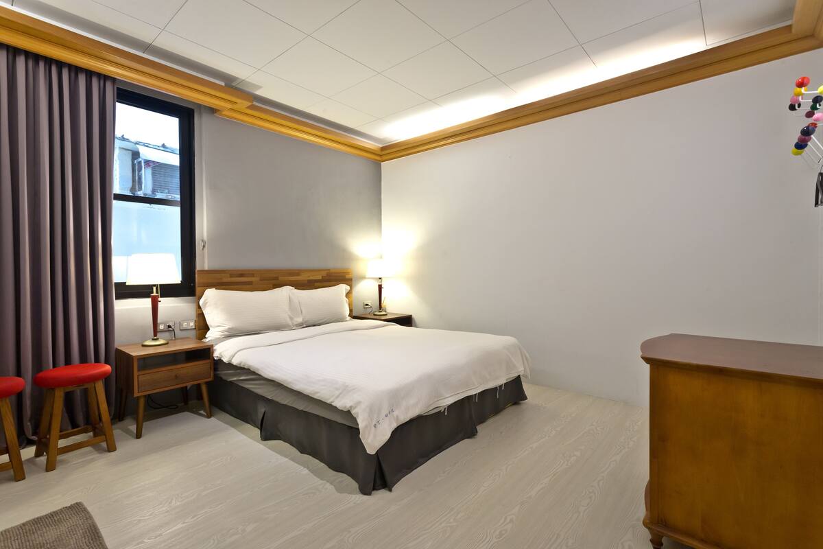 A hotel room with a bed, two red stools, a nightstand with a lamp, and a wooden dresser.