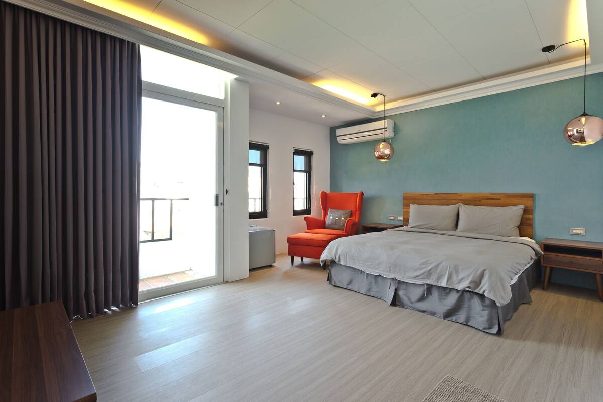 A modern bedroom with a large bed, a red chair, and a view of the outdoors through a sliding door.