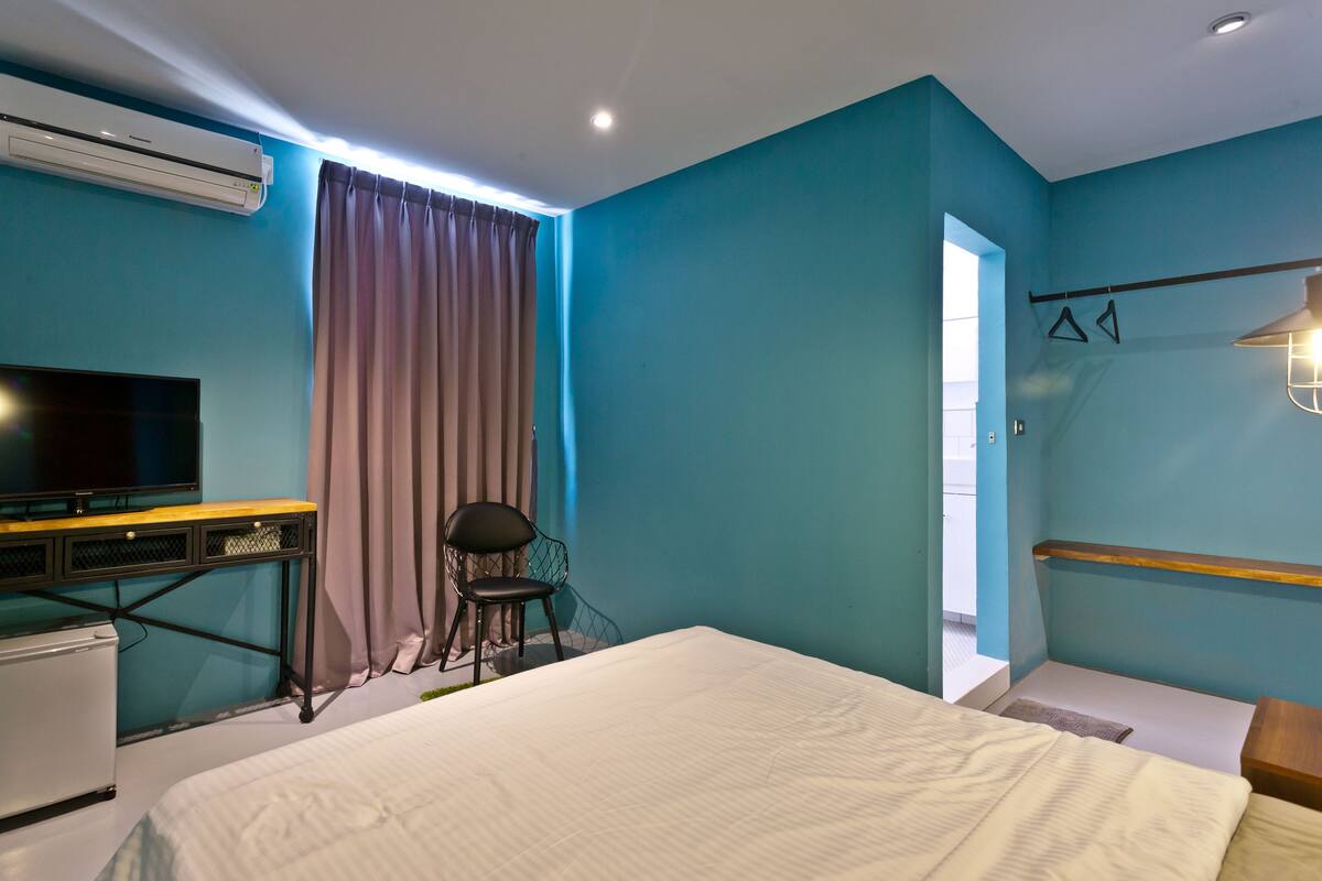 A bedroom with a bed, a desk, a chair, an air conditioner, and a television.