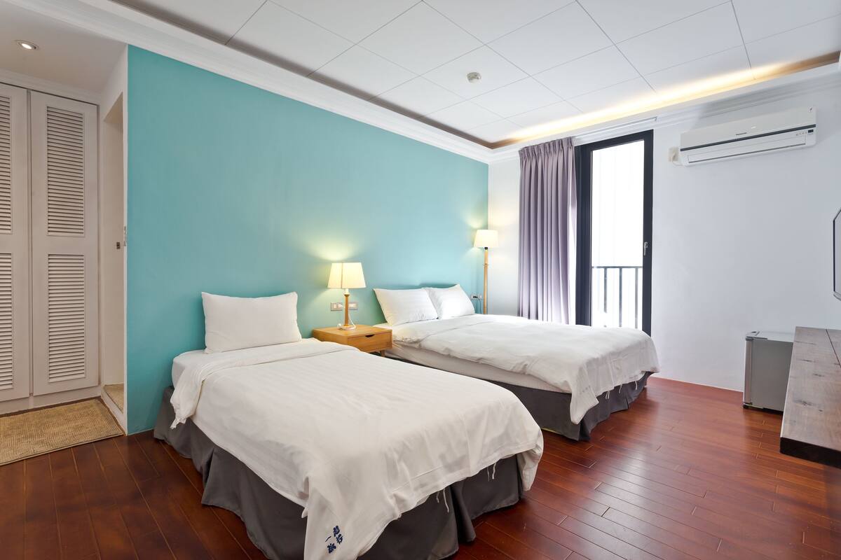 A hotel room with two beds, a teal accent wall, wooden flooring, a window with curtains, and an air conditioning unit.