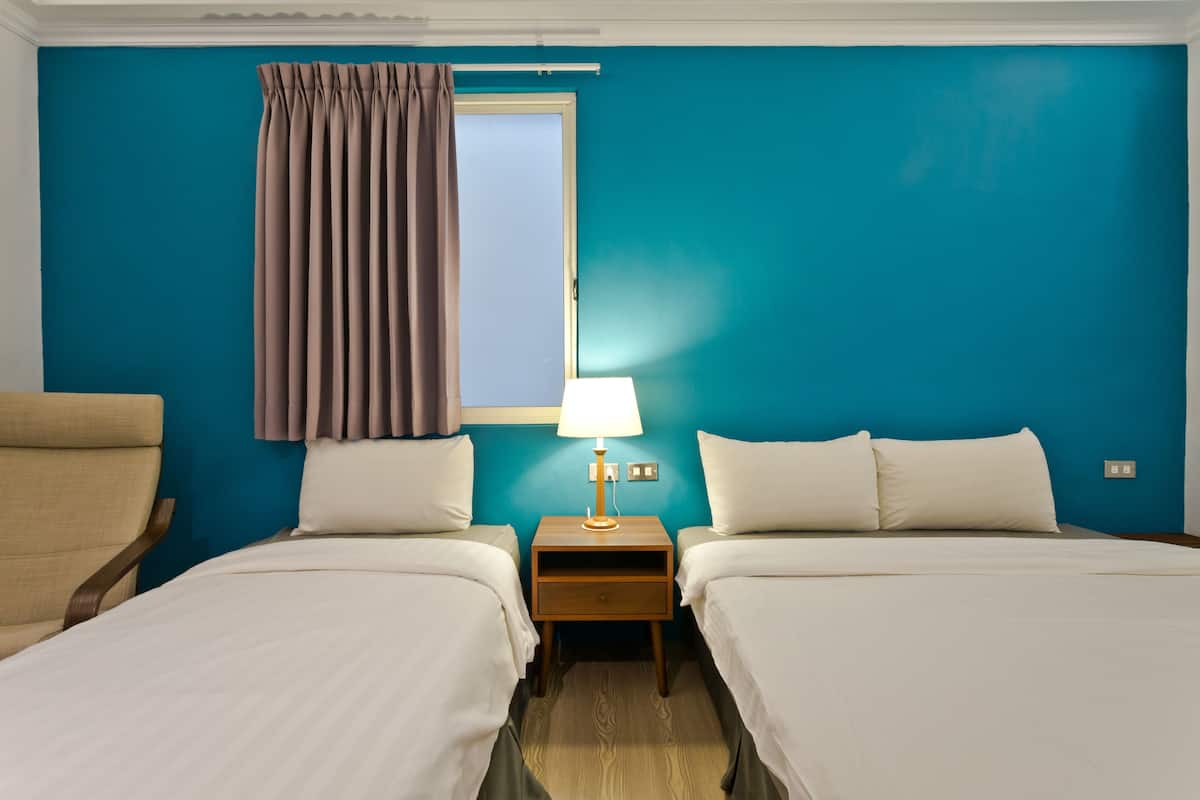 A hotel room with two beds, a nightstand with a lamp, a window with curtains, and a chair.