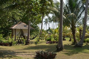Una cabaña con techo de paja, una hamaca y una mesa, rodeada de palmeras y plantas tropicales.