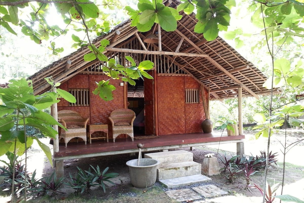 A rustic wooden house with a thatched roof, a porch with wicker furniture, and a small garden area.
