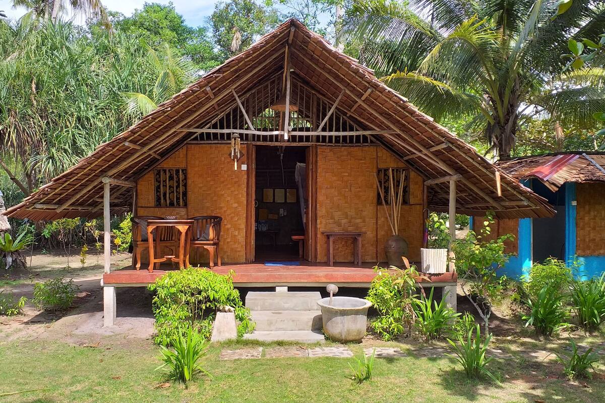 A rustic wooden house with a thatched roof, a porch with two wooden chairs, and a concrete water feature in front.