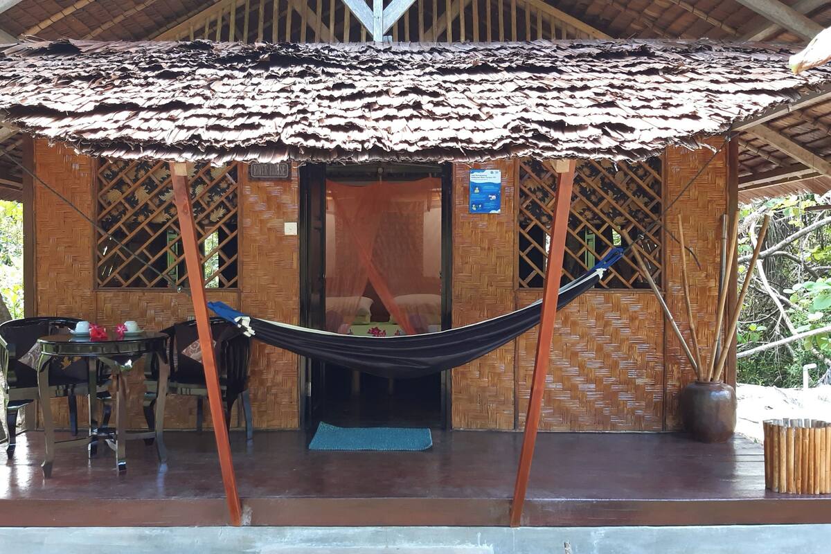 A rustic hut with a thatched roof, a hammock, and a table with a chair outside.