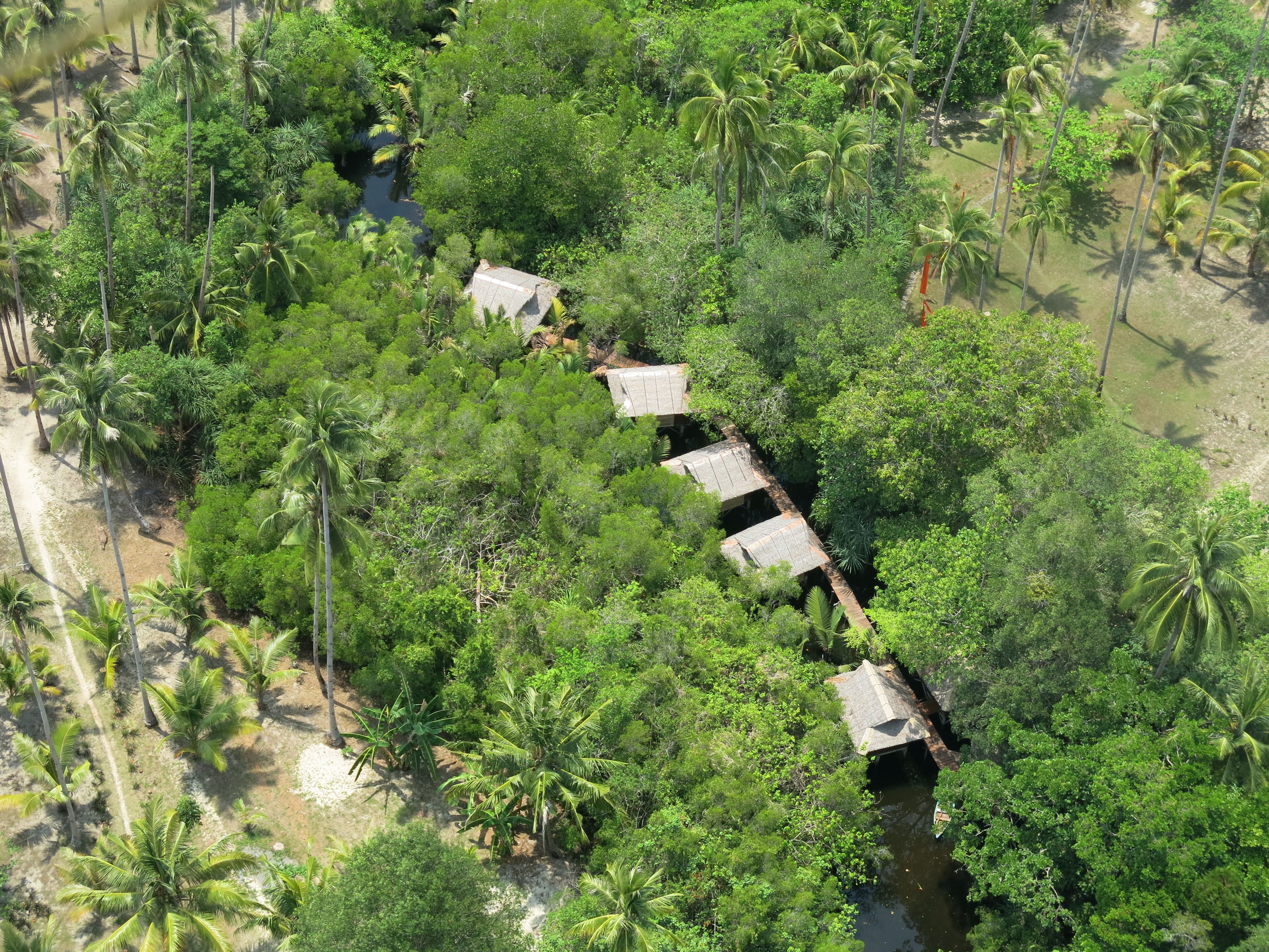 An aerial view of a remote, lush green area with a series of thatched-roof huts aligned along a narrow waterway.