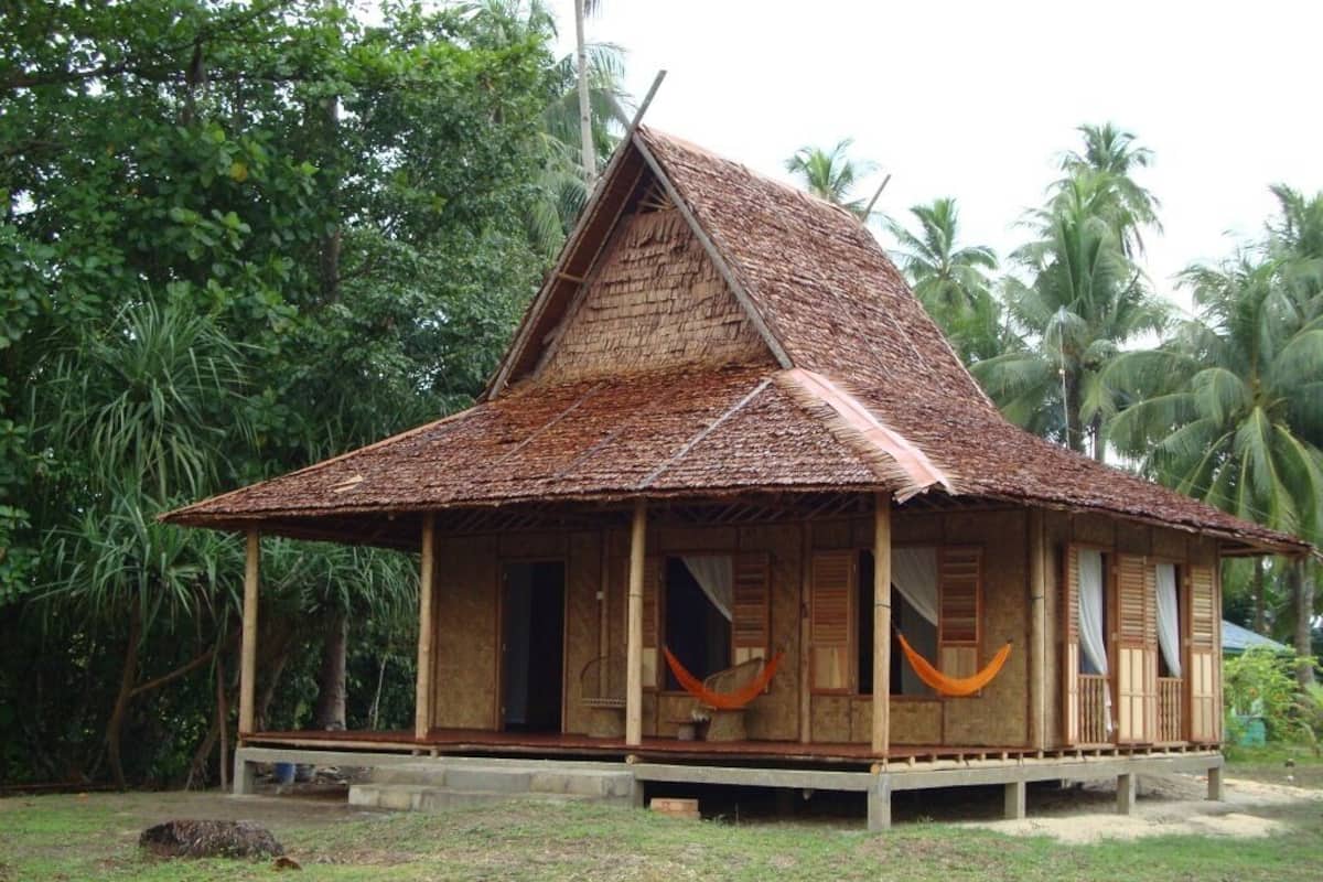 A wooden house with a thatched roof, surrounded by lush greenery.