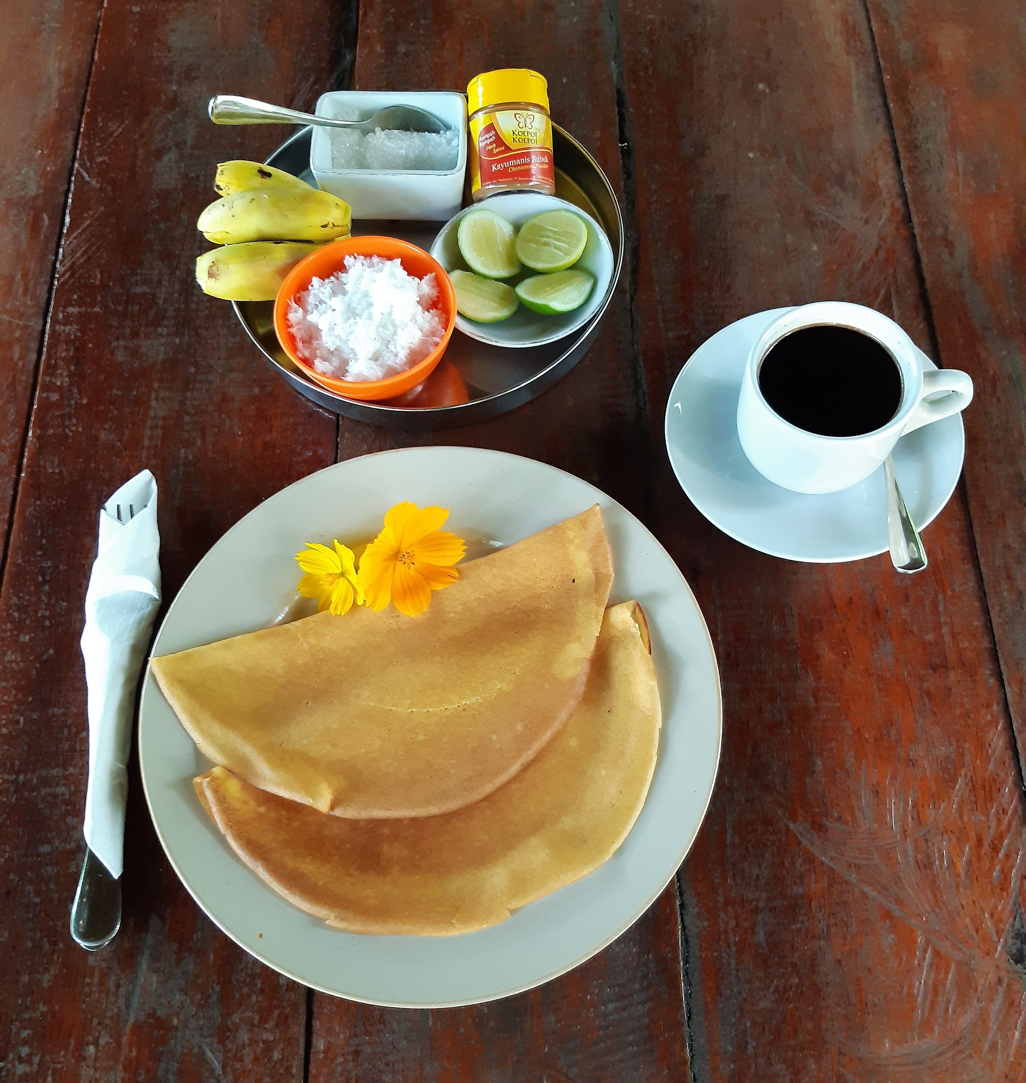 A plate of crepes with a flower, a bowl of sugar, a bowl of lime, a cup of coffee, and a container of salt on a wooden table.