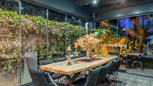 A modern dining area with a glass wall, a wooden table, black wireframe chairs, and a variety of plants.