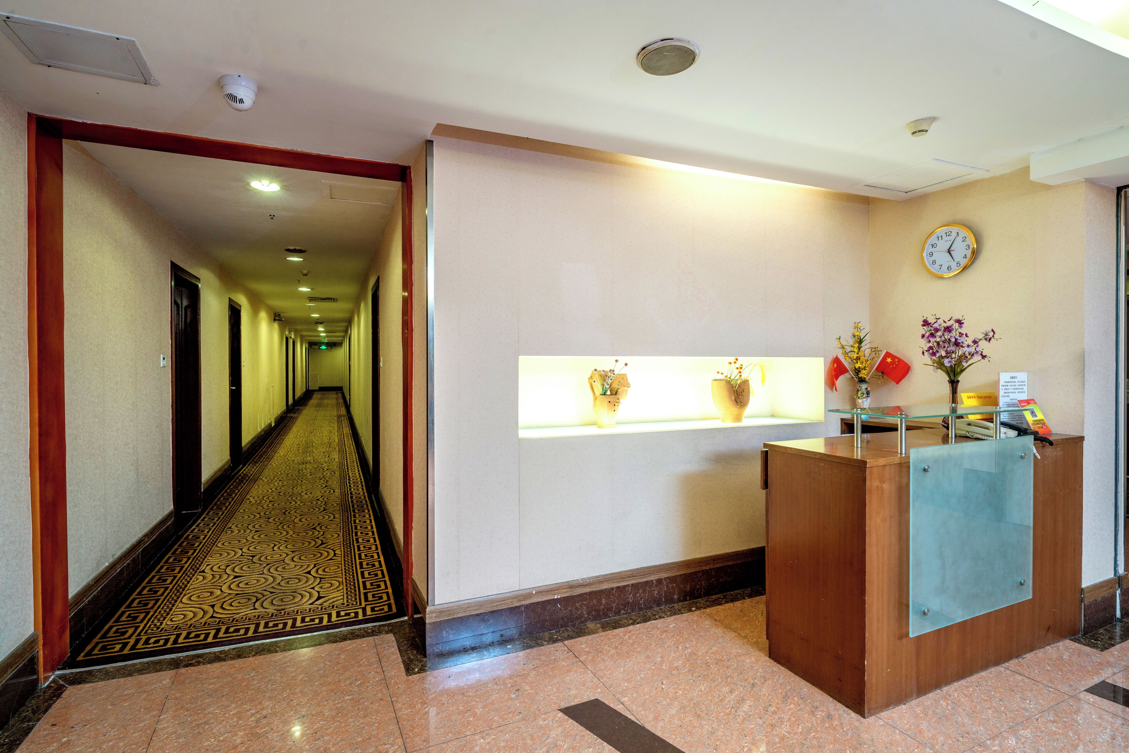 A hotel reception area with a reception desk, a clock on the wall, and a hallway leading to rooms.
