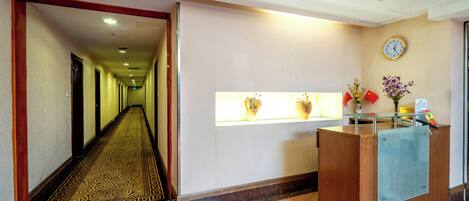 A hotel reception area with a reception desk, a clock on the wall, and a hallway leading to rooms.