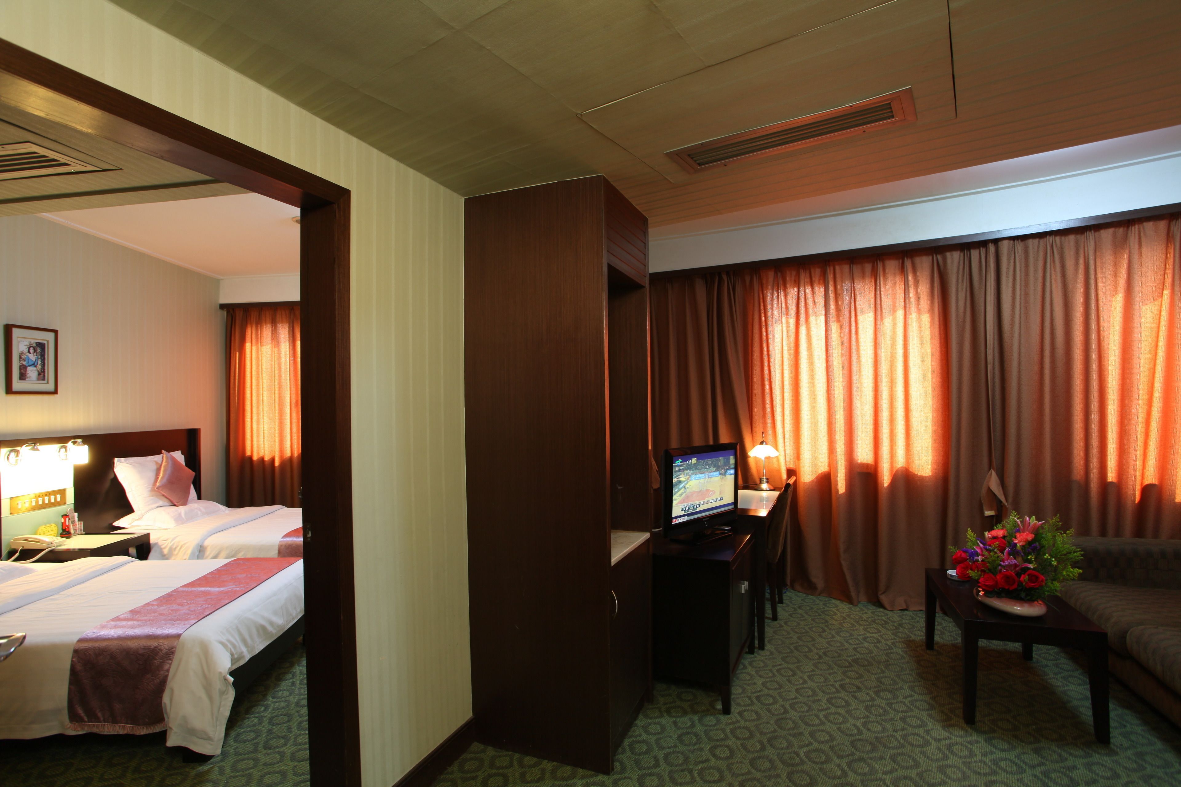 A hotel room with two beds, a desk with a television, a sofa, and a dining table with a flower arrangement.
