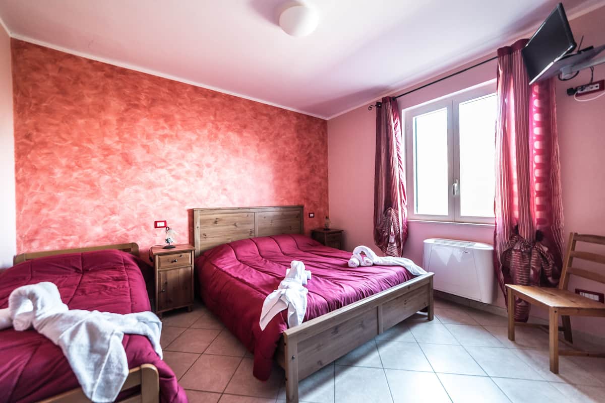 A bedroom with two beds, a wooden headboard, a nightstand, a window with curtains, a radiator, and a TV mounted on the wall.