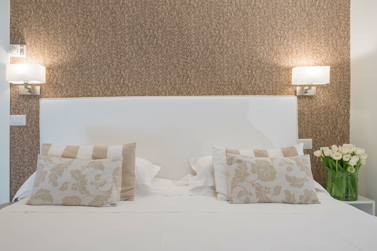 Una cama bien hecha con dos almohadones decorativos, una lámpara de pared y un florero con flores sobre una mesita de noche.