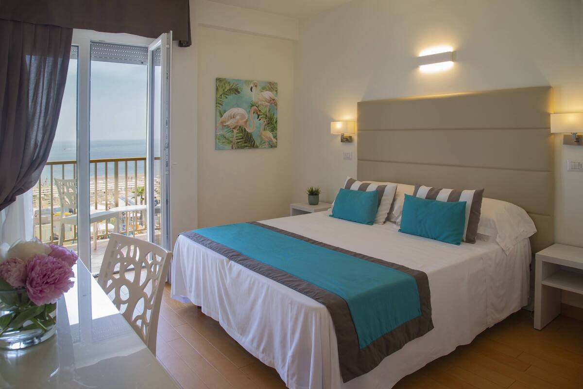 Un dormitorio con cama, mesita de noche, una silla, una mesa con un florero y un cuadro de flamencos, y vista a la playa desde una puerta abierta.
