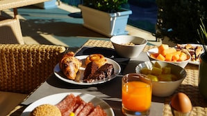 Una mesa de desayuno con diversos platos, un vaso de jugo de naranja y vistas a una piscina y follaje.
