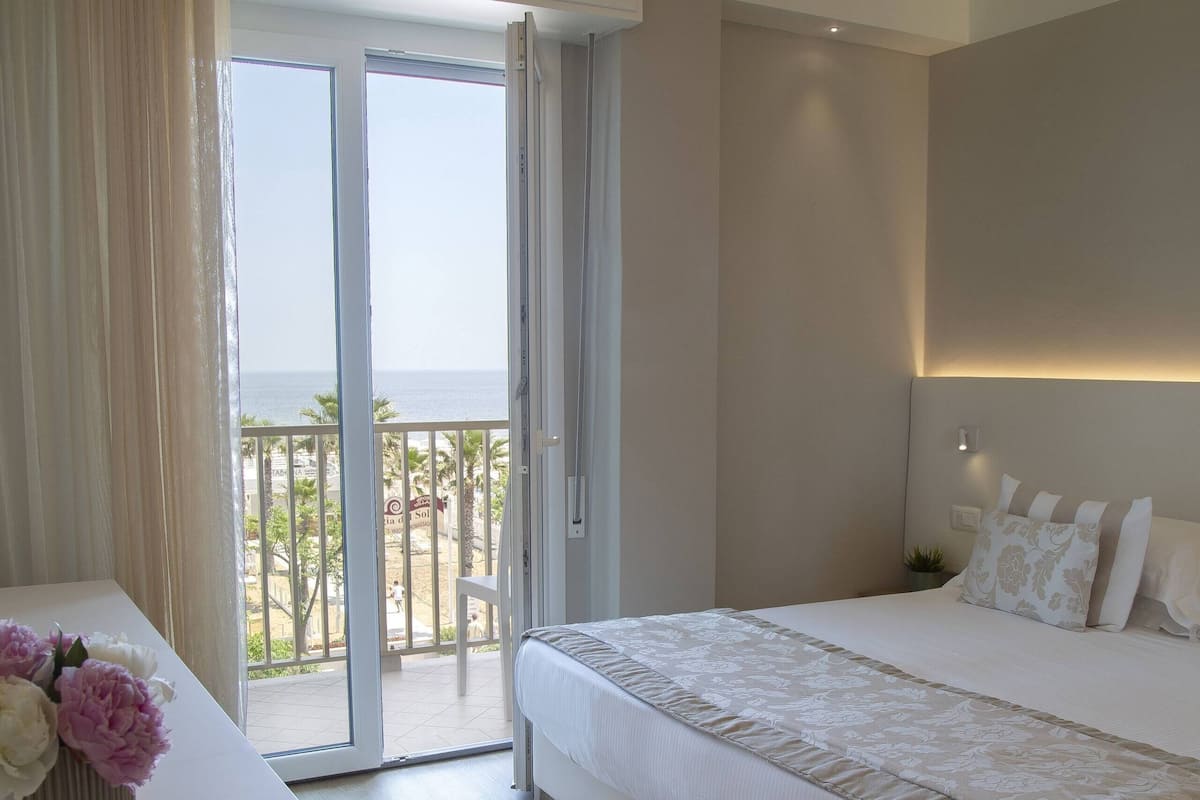 Habitación de hotel con una cama grande, una mesita de noche con un florero, un balcón con vistas a la playa y una puerta corrediza de cristal.