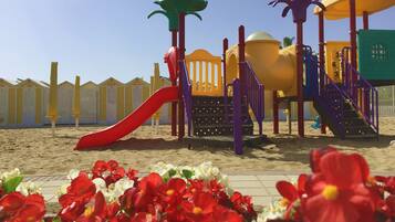 Un parque infantil con un tobogán rojo y diversas estructuras de juego, rodeado de arena y una fila de cabañas de playa al fondo.