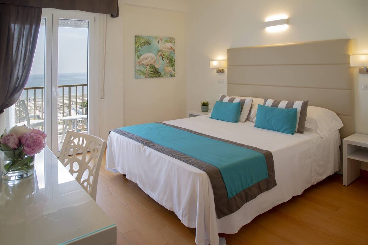 Un dormitorio con una cama grande, una mesita de noche, una silla, una mesa con un florero y vistas al mar a través de una puerta de balcón.