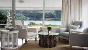 Quarto com vista para um lago e colinas, com duas cadeiras, uma mesa redonda e uma mesinha lateral.