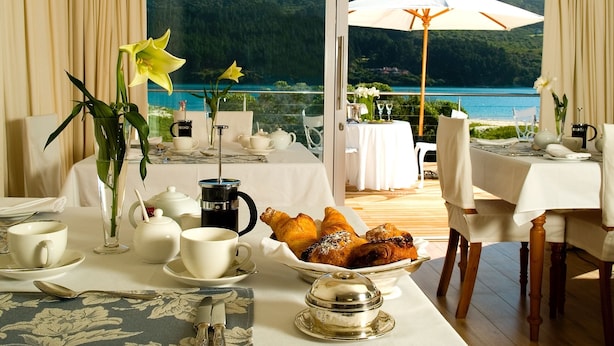 Área de refeições com mesas preparadas para o café da manhã, com vista para um lago e colinas panorâmicos.