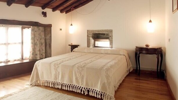 A bedroom with a large bed, wooden flooring, a stone fireplace, and a window with curtains.