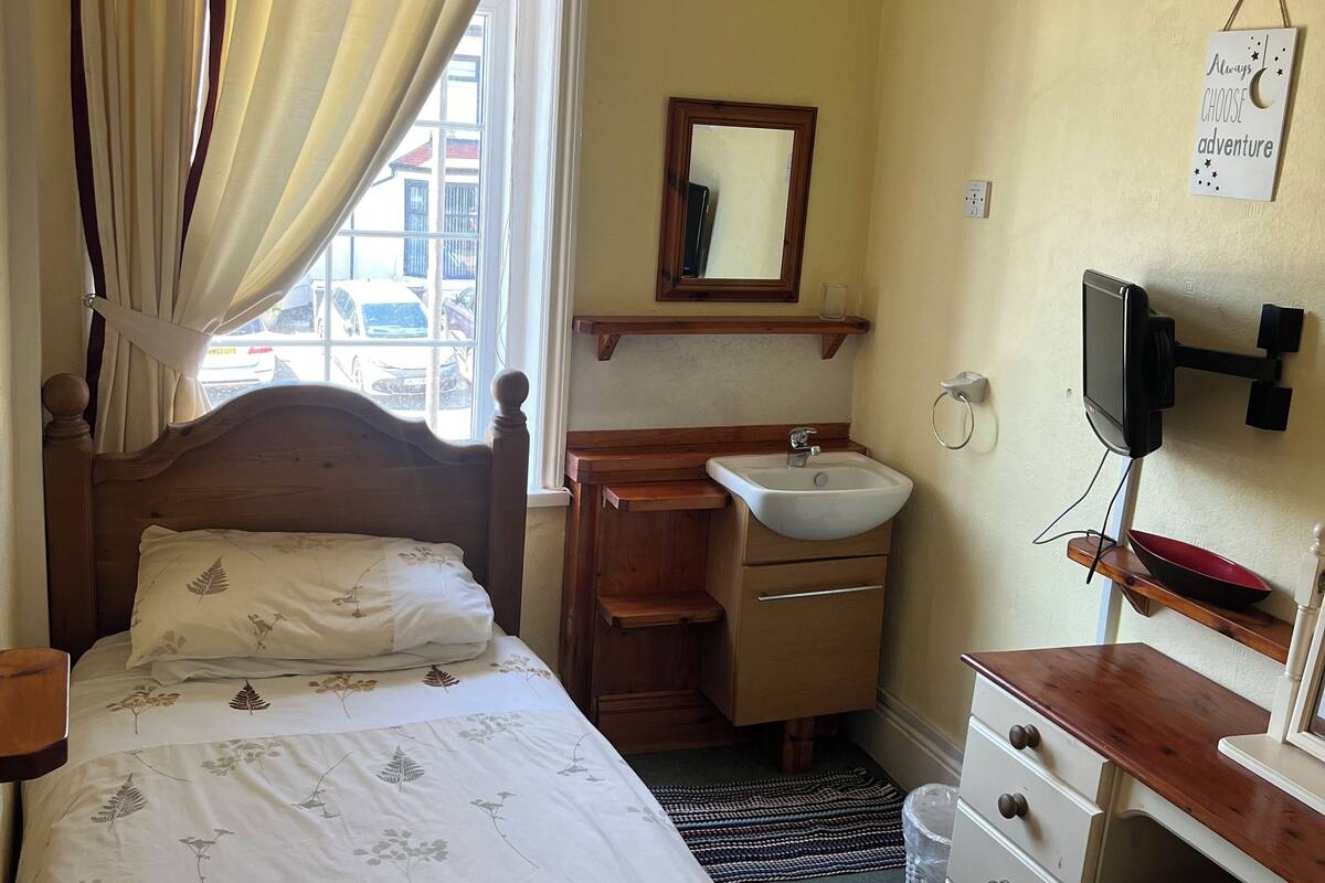 A bedroom with a bed, a bedside table, a wooden dresser, a mirror, a sink, and a window with curtains.