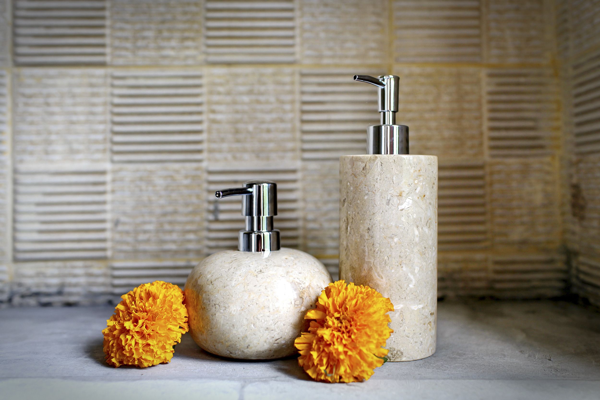 Two cylindrical containers with pumps, placed on a flat surface with two marigold flowers beside them.
