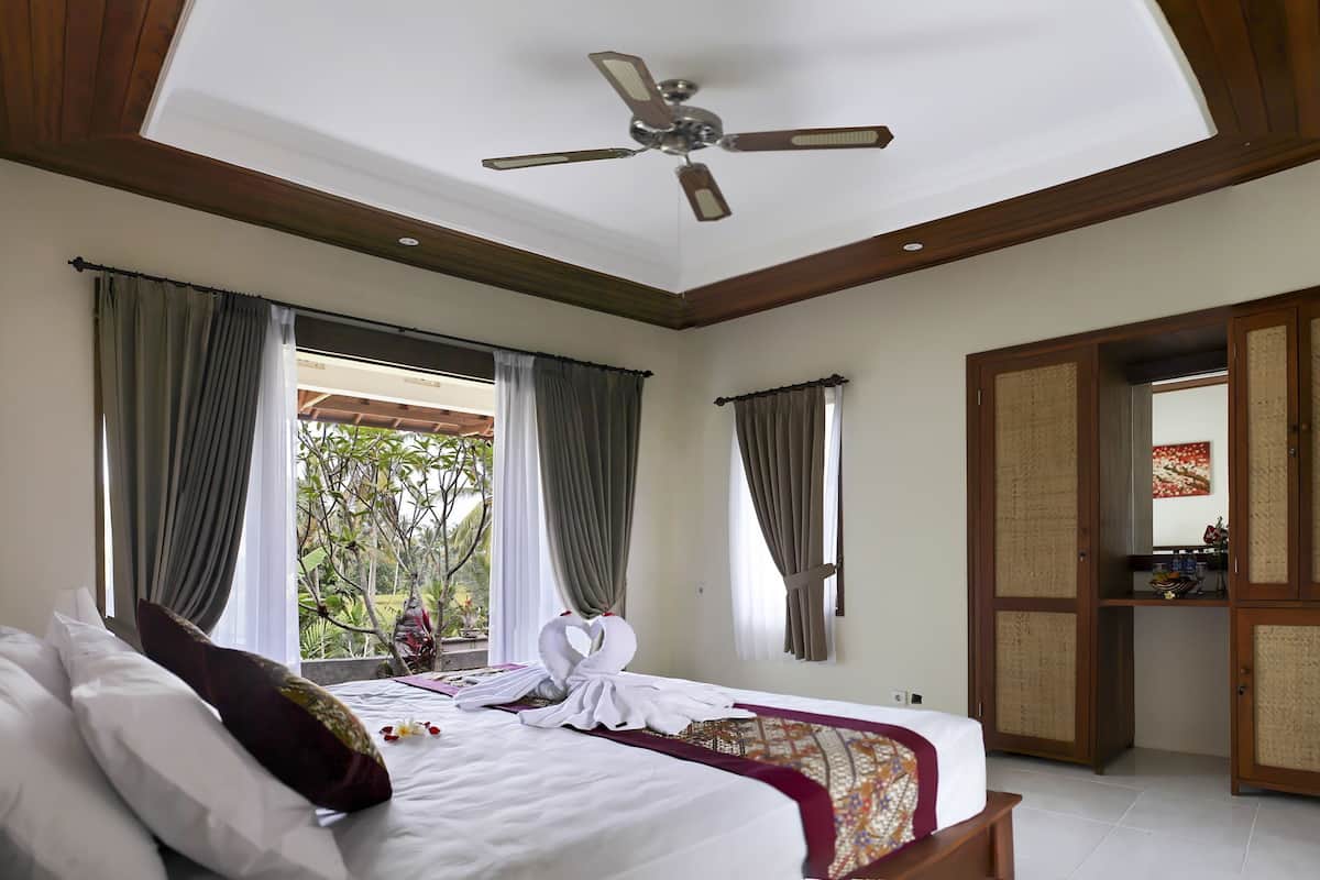 Habitación de hotel con una cama grande, ventilador de techo y vistas a zonas verdes.
