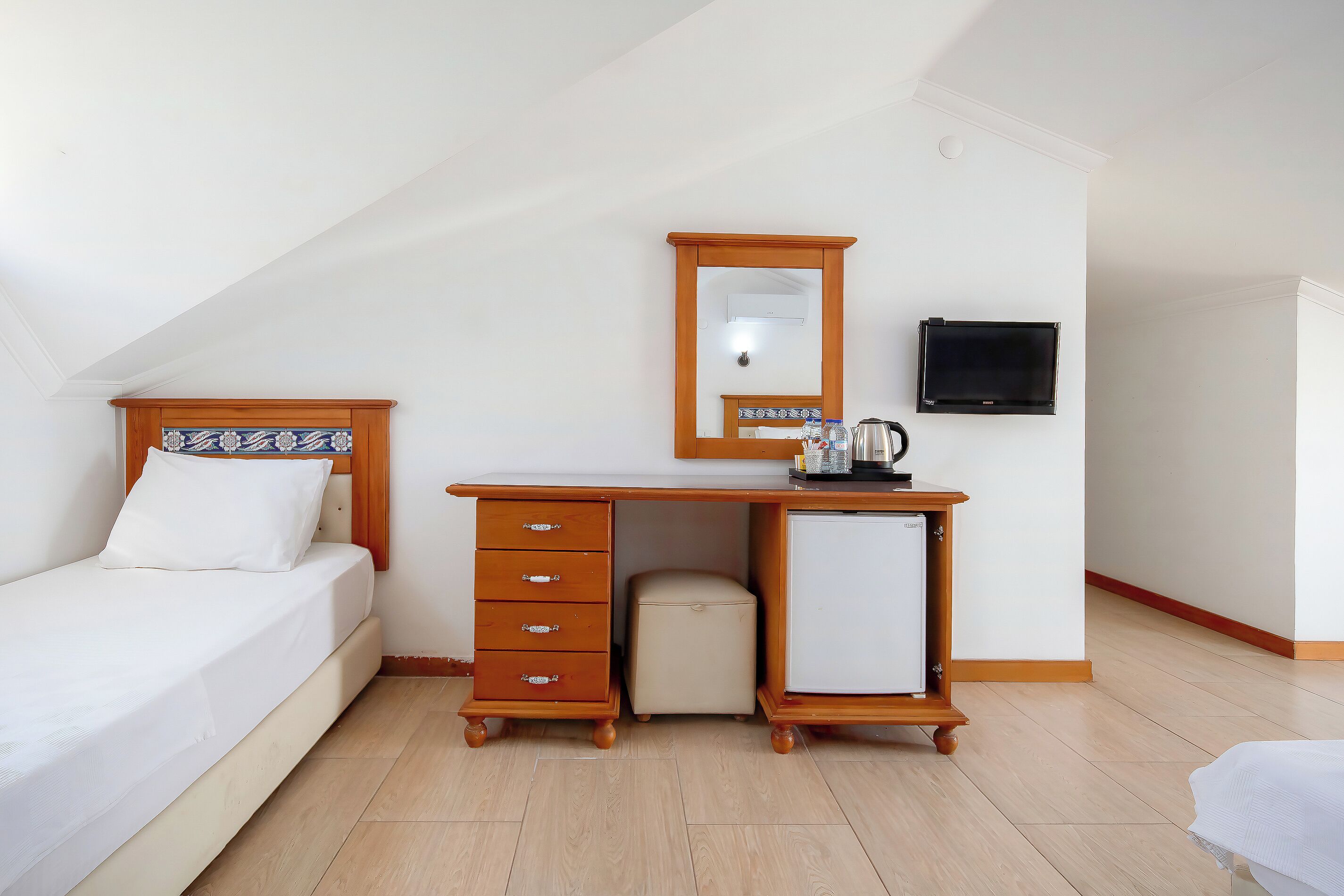 Een kamer met een bed, een houten bureau met lades, een kleine koelkast, een spiegel en een televisie aan de muur.