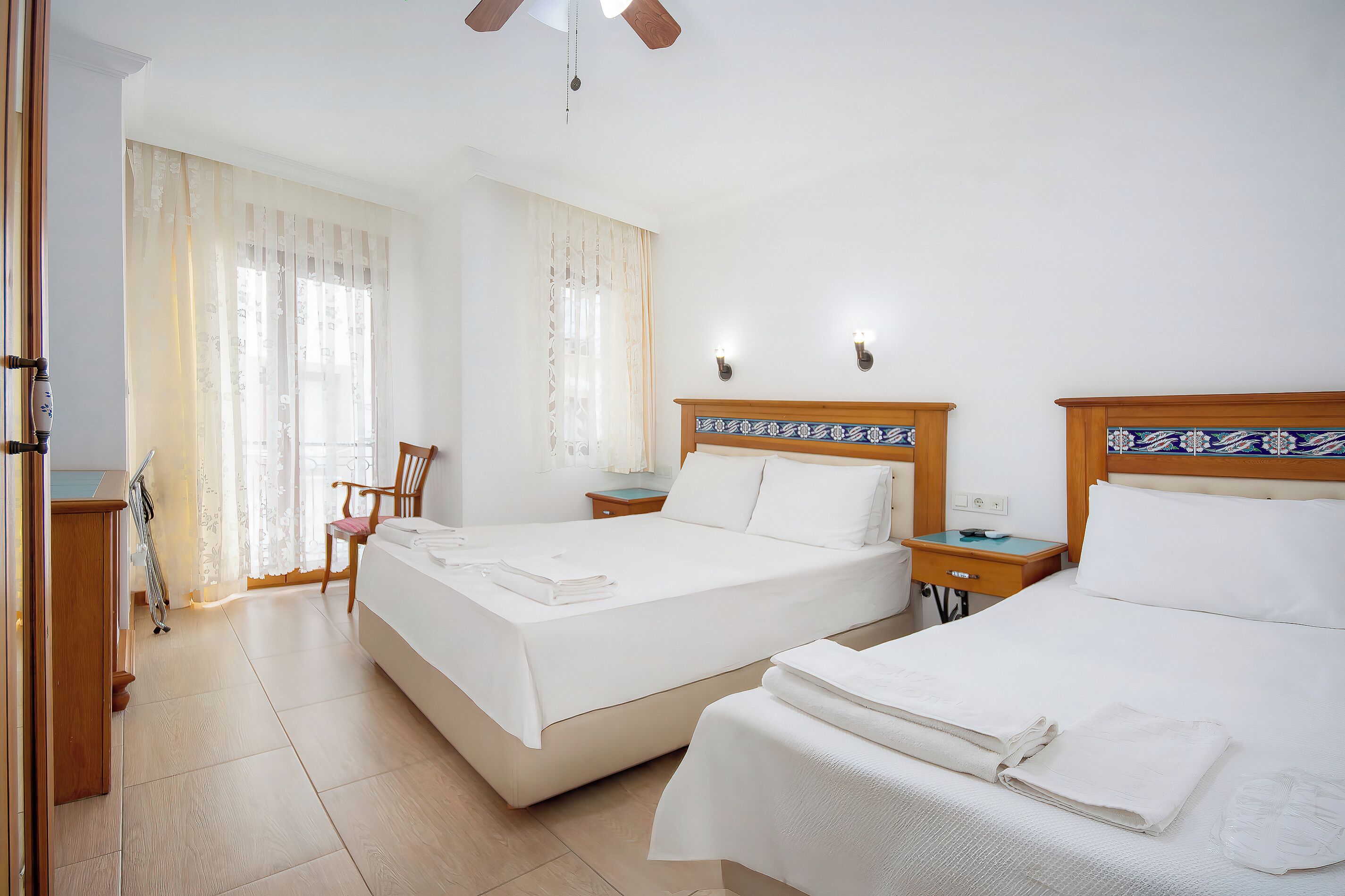 A hotel room with two beds, a wooden headboard, a nightstand, a chair, and a window with sheer curtains.
