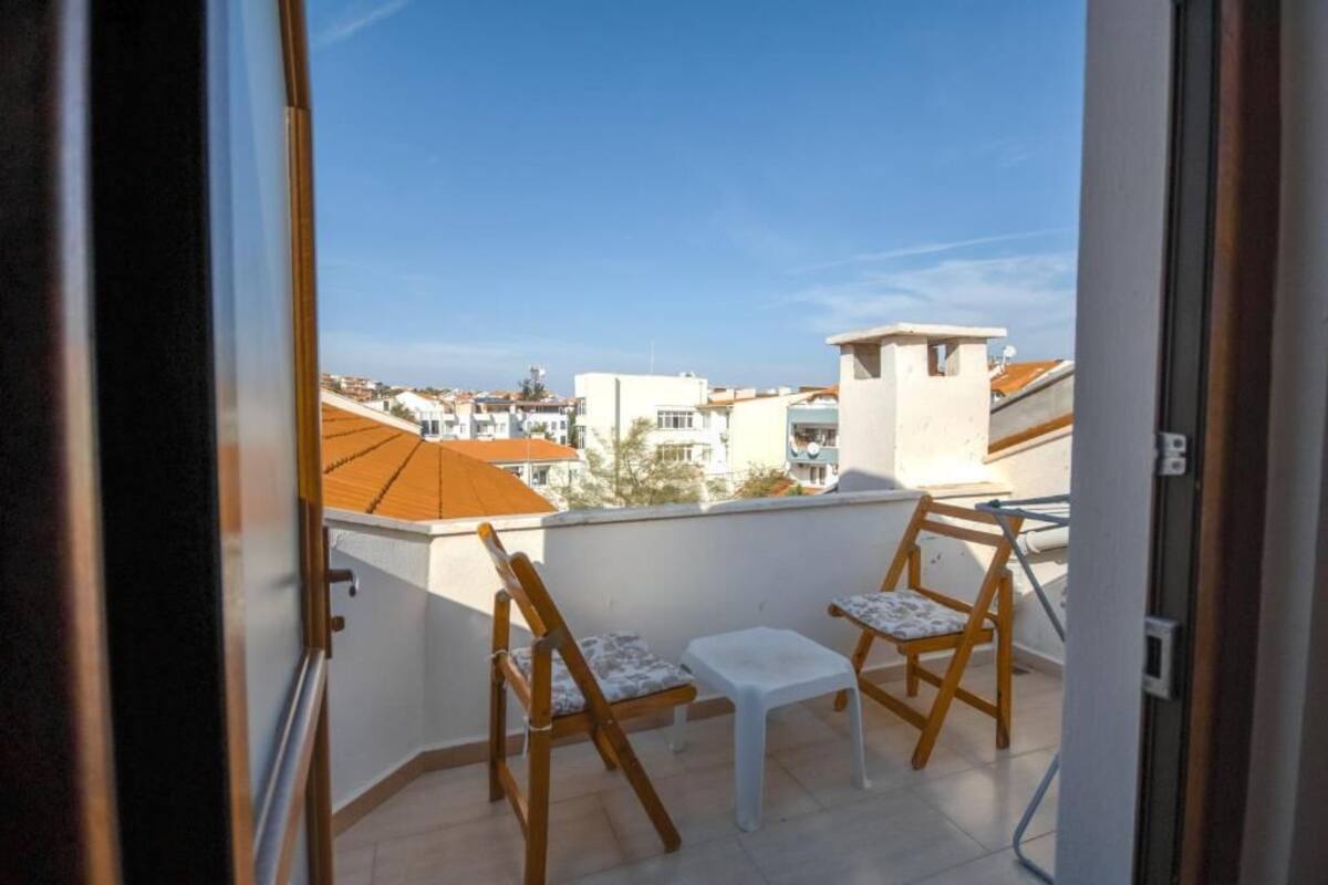 A balcony with two wooden chairs and a small table, overlooking a residential area with orange-roofed buildings.