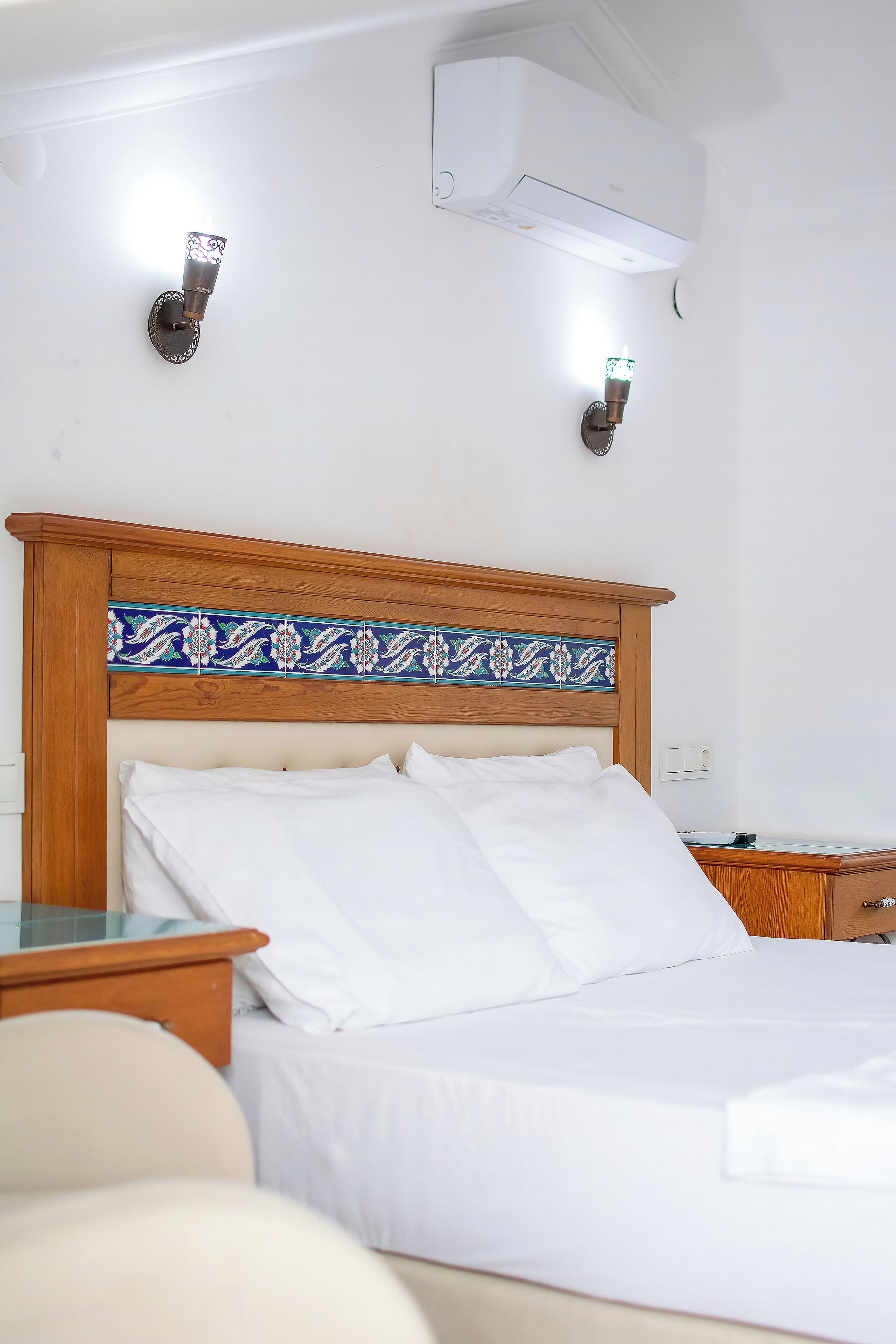 A neatly made bed with white linens, a wooden headboard with a decorative tile pattern, and two wall-mounted light fixtures.