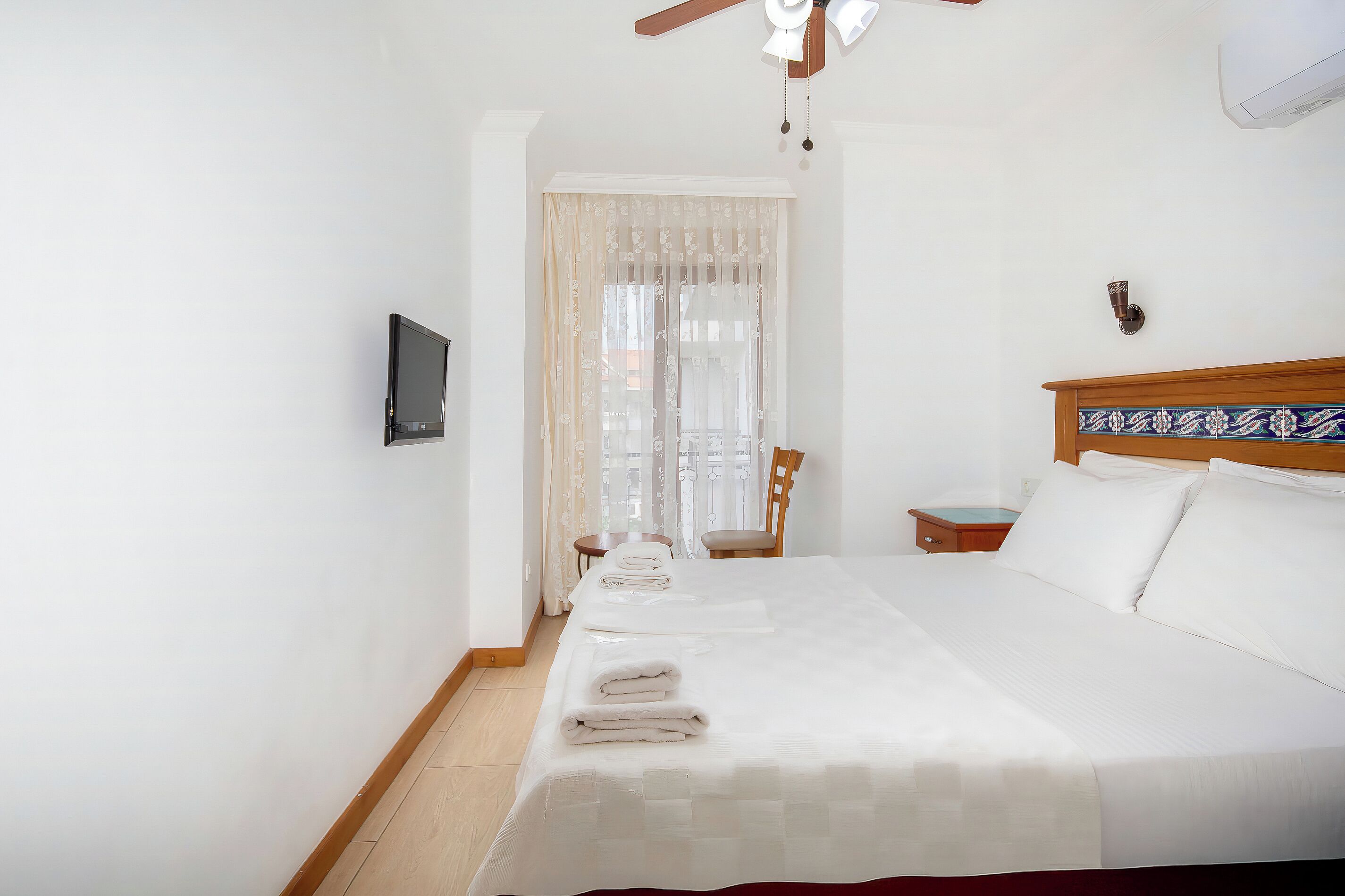 Un lit bien fait, avec du linge de lit blanc et une couvre-lit rouge, une table de chevet en bois avec une lampe, une chaise et un téléviseur fixé au mur.