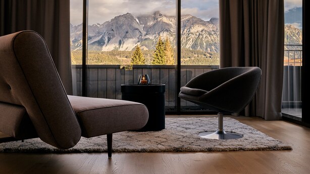 Una sala de estar moderna con un sofá y un sillón, una mesita con una vela, y vistas a montañas a través de amplios ventanales.