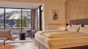 Habitación de hotel con una cama grande, una silla, una mesita y vistas a las montañas a través de un ventanal.