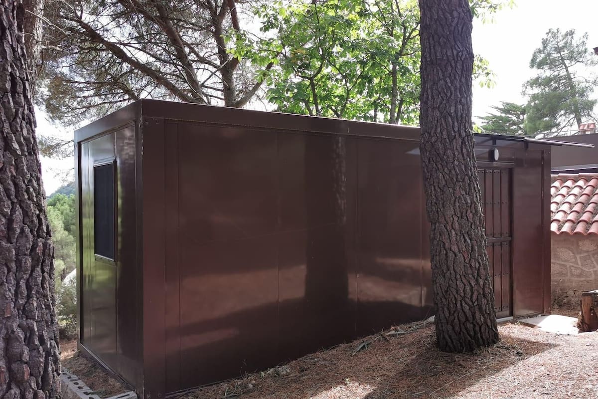 Una pequeña construcción de madera oscura, con una puerta y una ventana, rodeada de árboles y un muro de piedra.