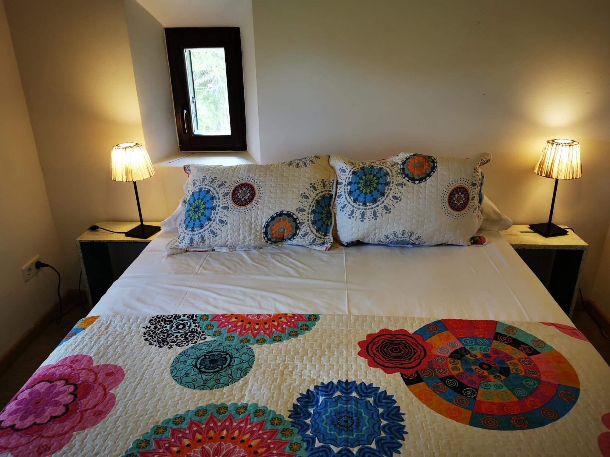 A bed with a quilt featuring colorful mandala patterns, two bedside lamps, and a window above the bed.
