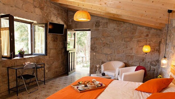 A cozy room with a stone wall, a bed with orange bedding, a dining table with a tray of food, and a chair.