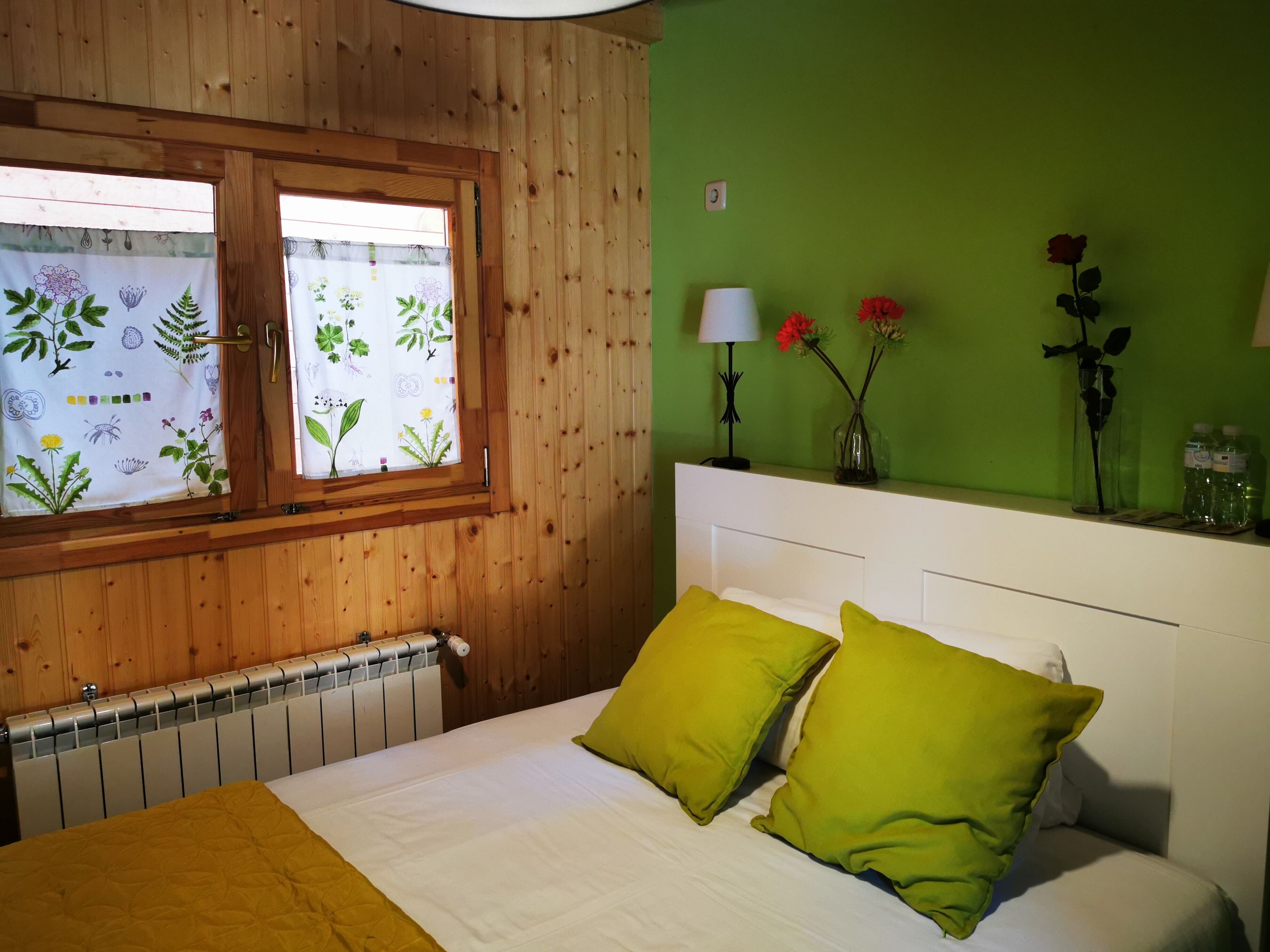 Una camera da letto con una testiera in legno, un letto con biancheria da letto bianca e cuscini verdi, un termosifone, una finestra con tende floreali, una lampada da comodino e un vaso con fiori su una mensola.