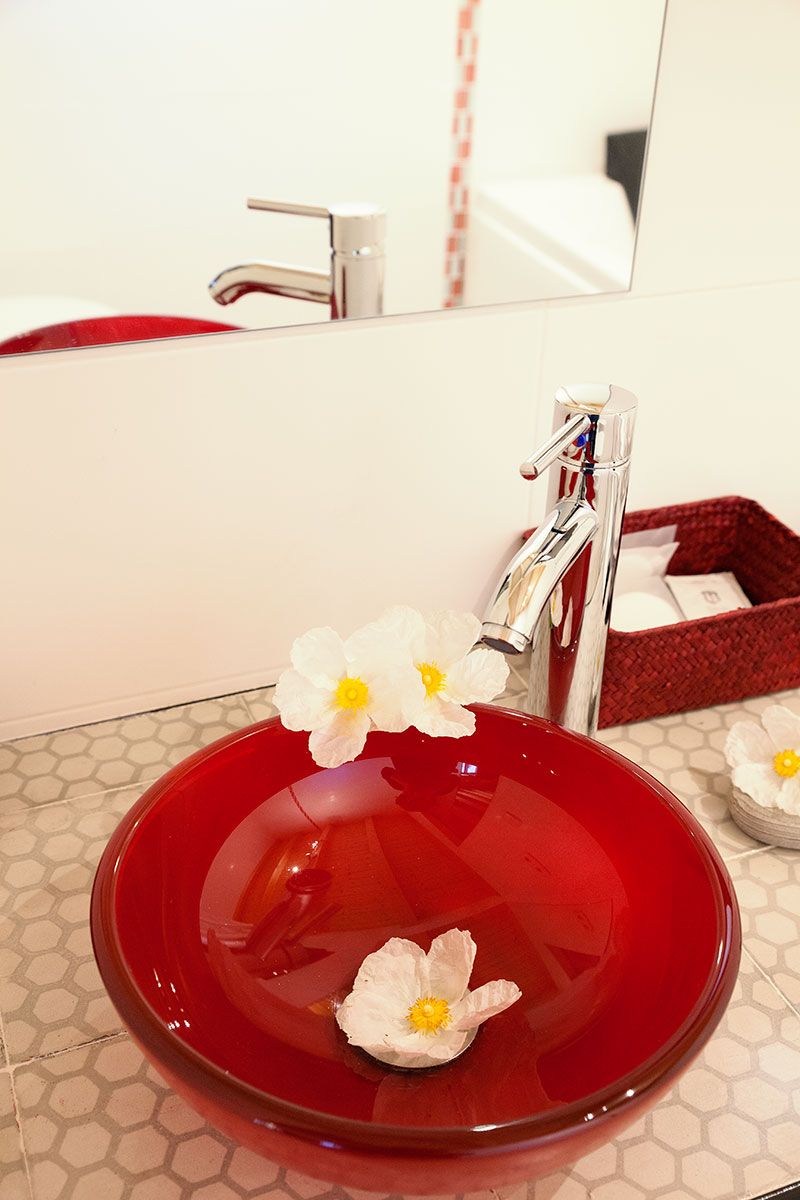 A red glass sink with a chrome faucet, white flowers, and a red basket with toiletries.