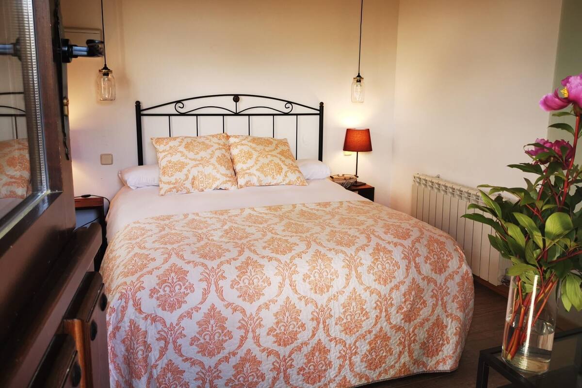 Una cama bien hecha con una colcha estampada, una lámpara de noche y una planta en una mesita de noche.