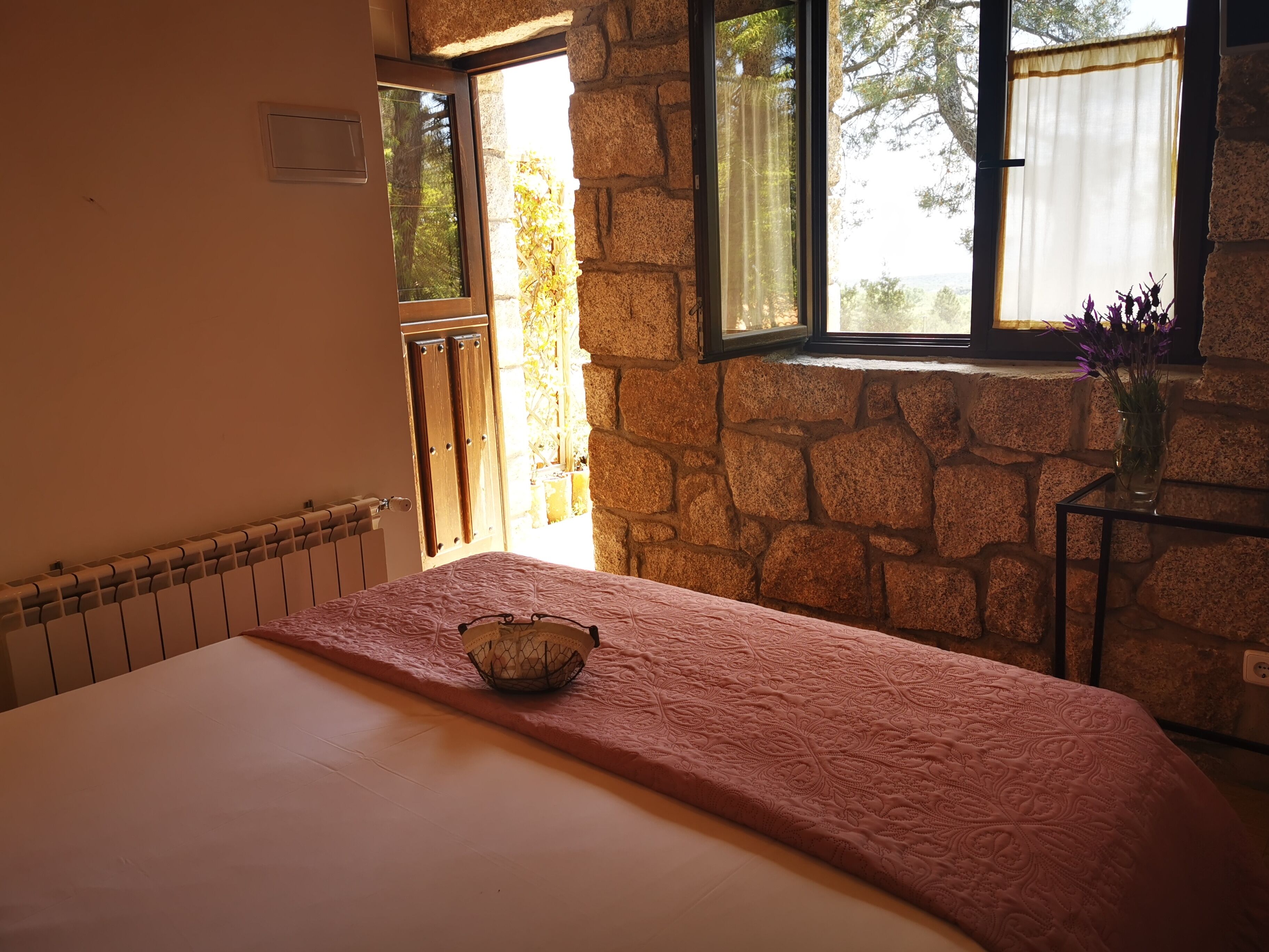 Un dormitorio con pared de piedra, una cama con colcha rosa, una ventana con cortina y un jarrón con flores sobre una mesita.
