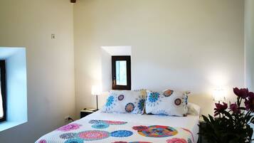 A bedroom with a wooden ceiling, a bed with a colorful quilt, bedside lamps, a window, and a vase with flowers.