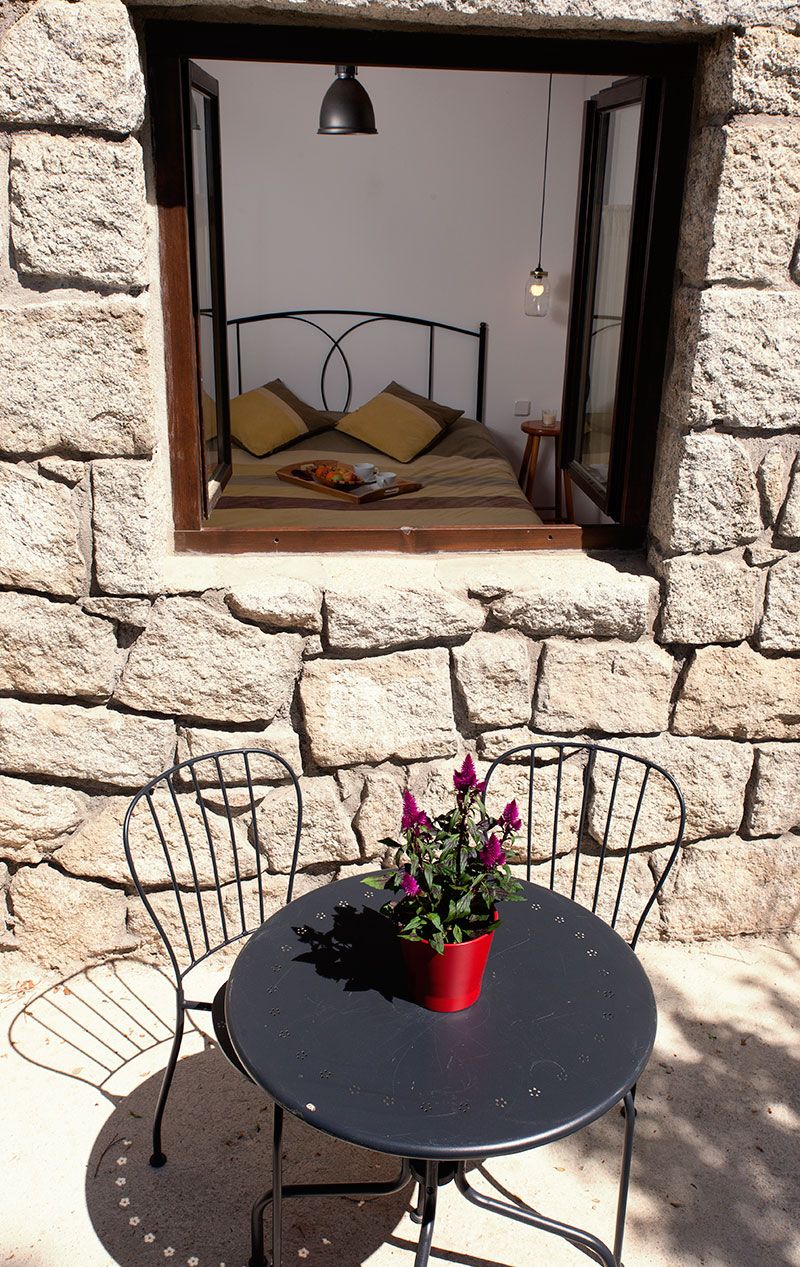 A stone wall with a window overlooking a bed with pillows and a bedside table. A round table with chairs and a potted plant is in the foreground.