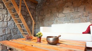A stone-walled room with a wooden ladder, a white couch, a wooden table with a metal bowl and a flower pot.