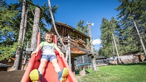Un niño resbala por un tobogán rojo en un área boscosa, con una cabaña de madera al fondo.